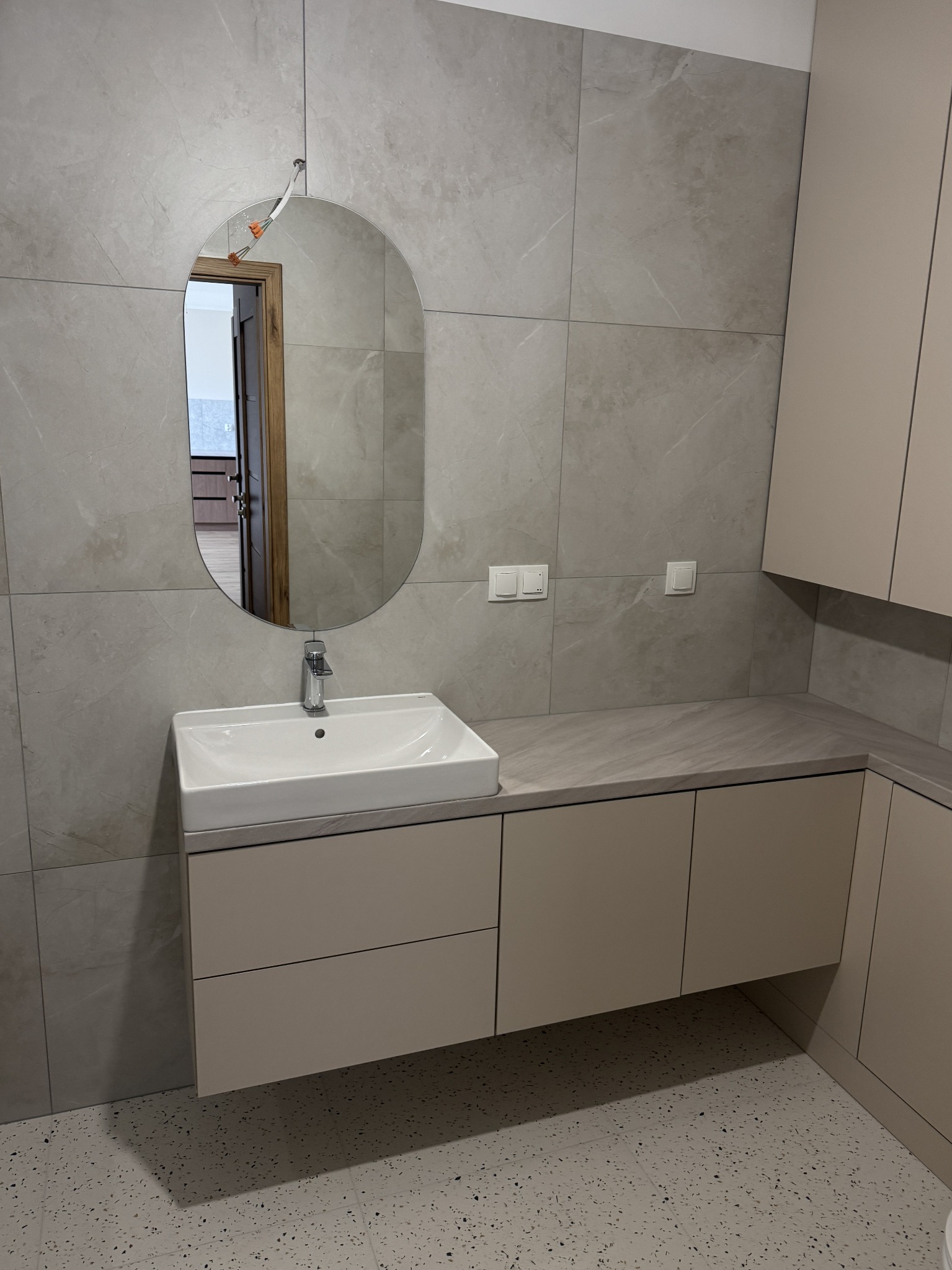 Wykończona łazienka z owalnym lustrem, beżowymi płytkami imitującymi marmur na ścianach i podłodze w stylu lastryko. Umywalka nablatowa z szafką w kolorze beżowym.