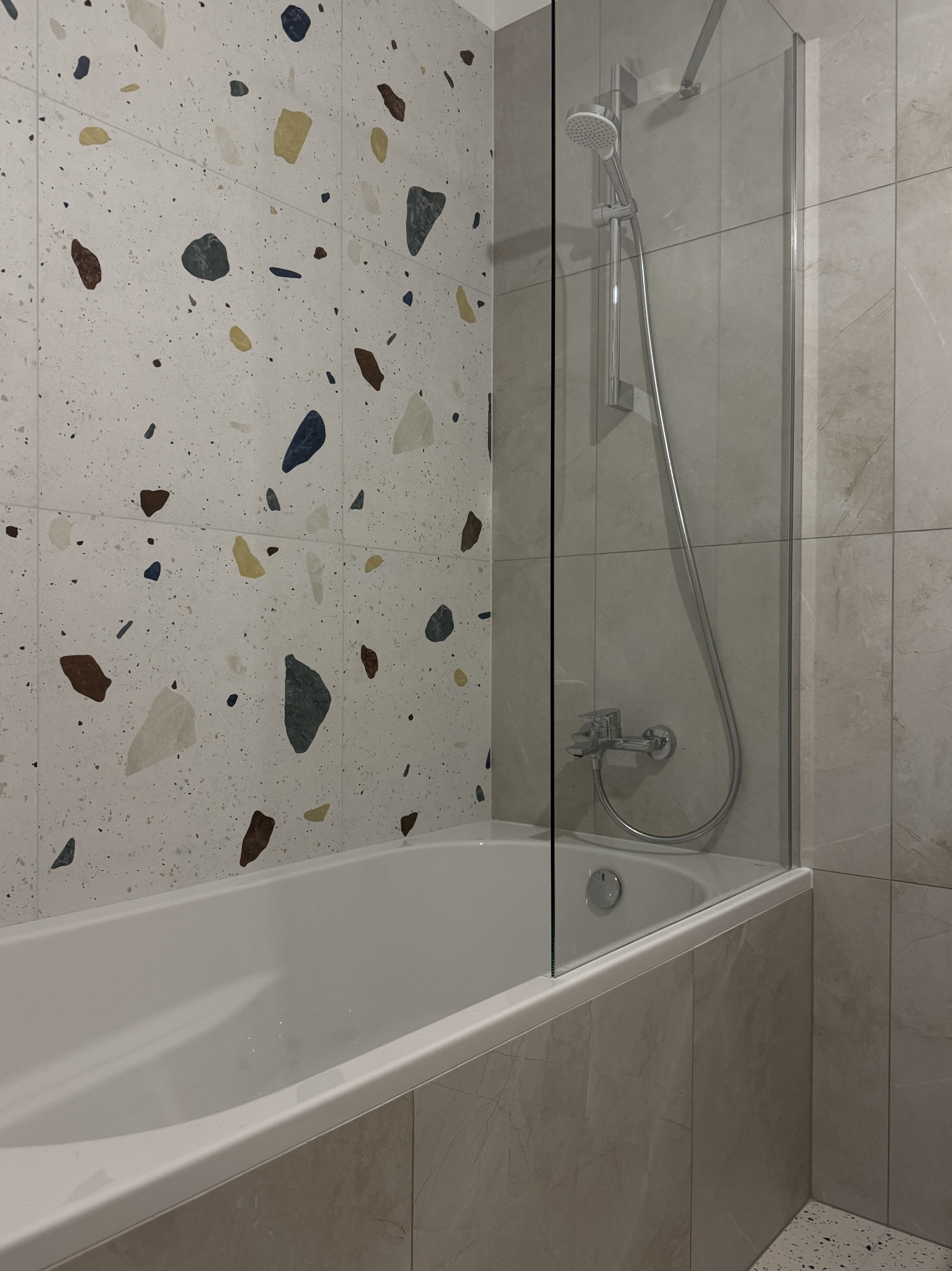 Wykończona łazienka z wanną, szklanym parawanem nawannowym i oryginalną mozaiką na ścianie. Widoczne baterie i płytki imitujące marmur.