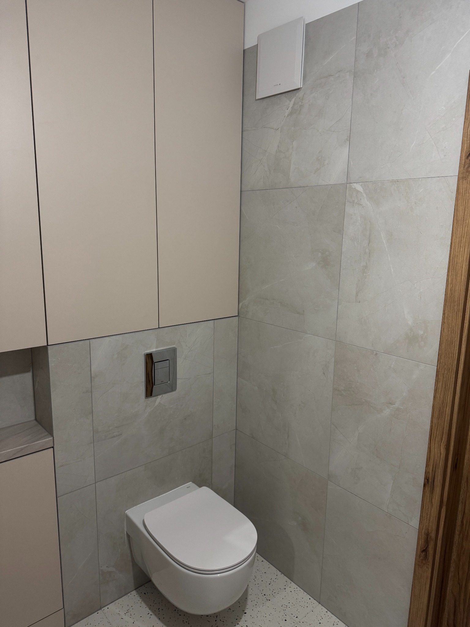 Nowoczesna toaleta w beżowo-szarej tonacji: podwieszana muszla, płytki imitujące marmur, zabudowa meblowa i dyskretny wentylator.