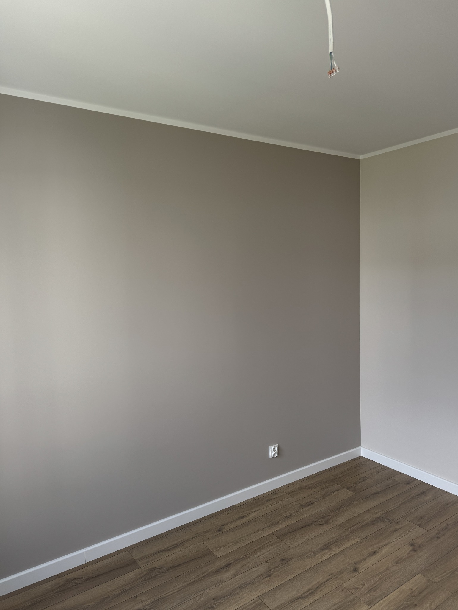 Pokój po malowaniu ścian na szaro, z drewnianą podłogą i białymi listwami przypodłogowymi. Widoczne gniazdko elektryczne i wystające kable z sufitu.