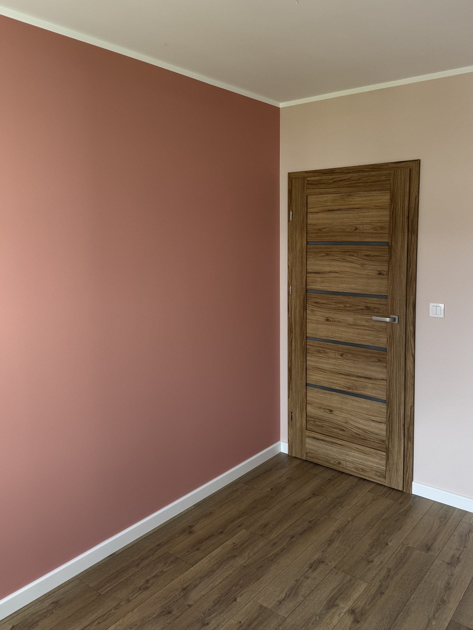 Pomieszczenie po malowaniu ścian na odcienie różu, z drewnianą podłogą i dopasowaną kolorystycznie stolarką drzwiową z poziomymi przeszkleniami.