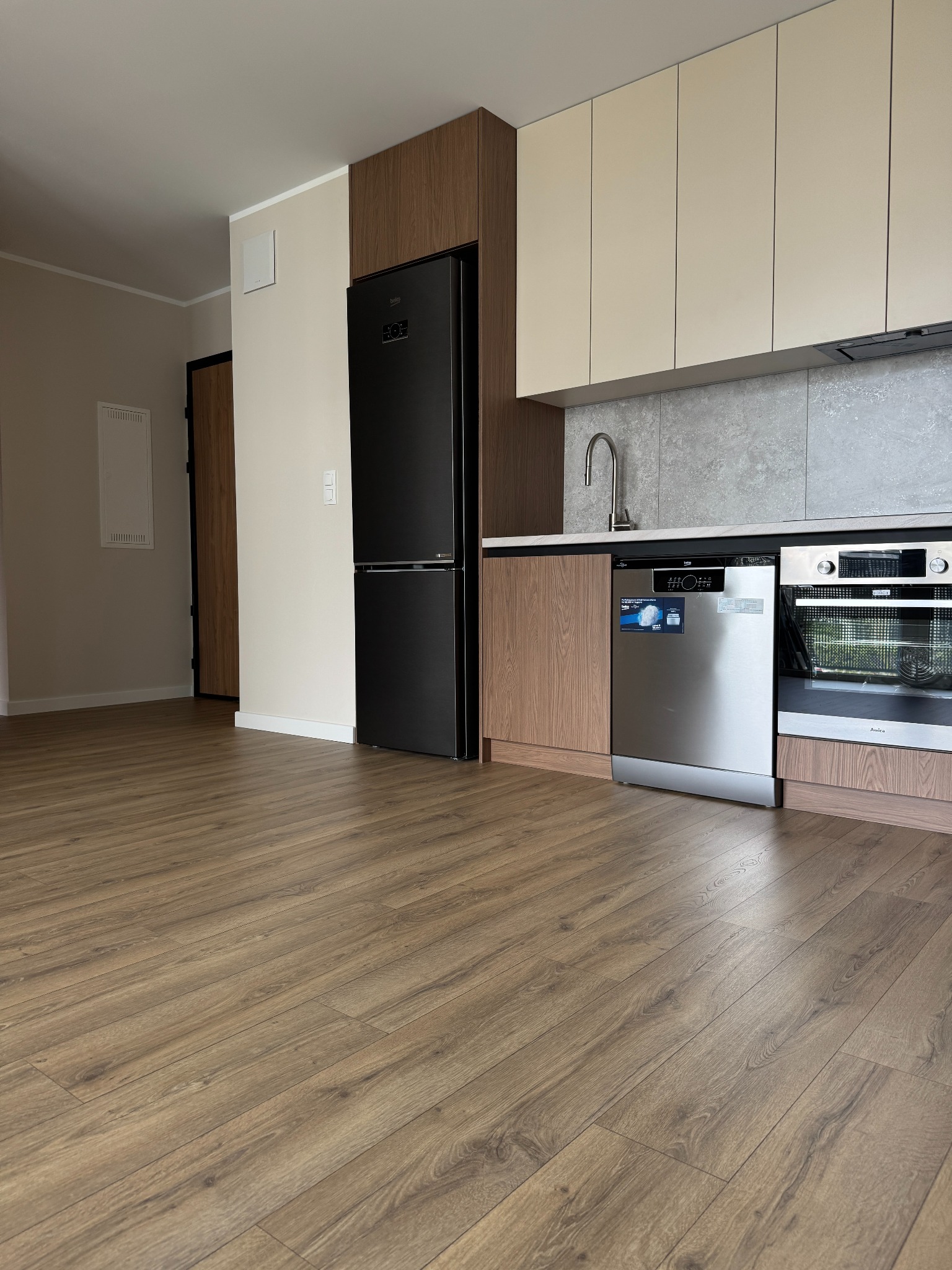 Jasna kuchnia z drewnianą podłogą, zabudową w beżu i brązie, sprzęt AGD w zabudowie: lodówka Beko, zmywarka i piekarnik Amica. Minimalistyczny design.