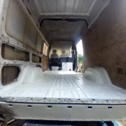 Wnętrze furgonetki w trakcie renowacji, widok z tyłu na odświeżoną, pomalowaną na biało podłogę, surowe ściany boczne bez tapicerki, widoczne ślady korozji i spawania na karoserii.