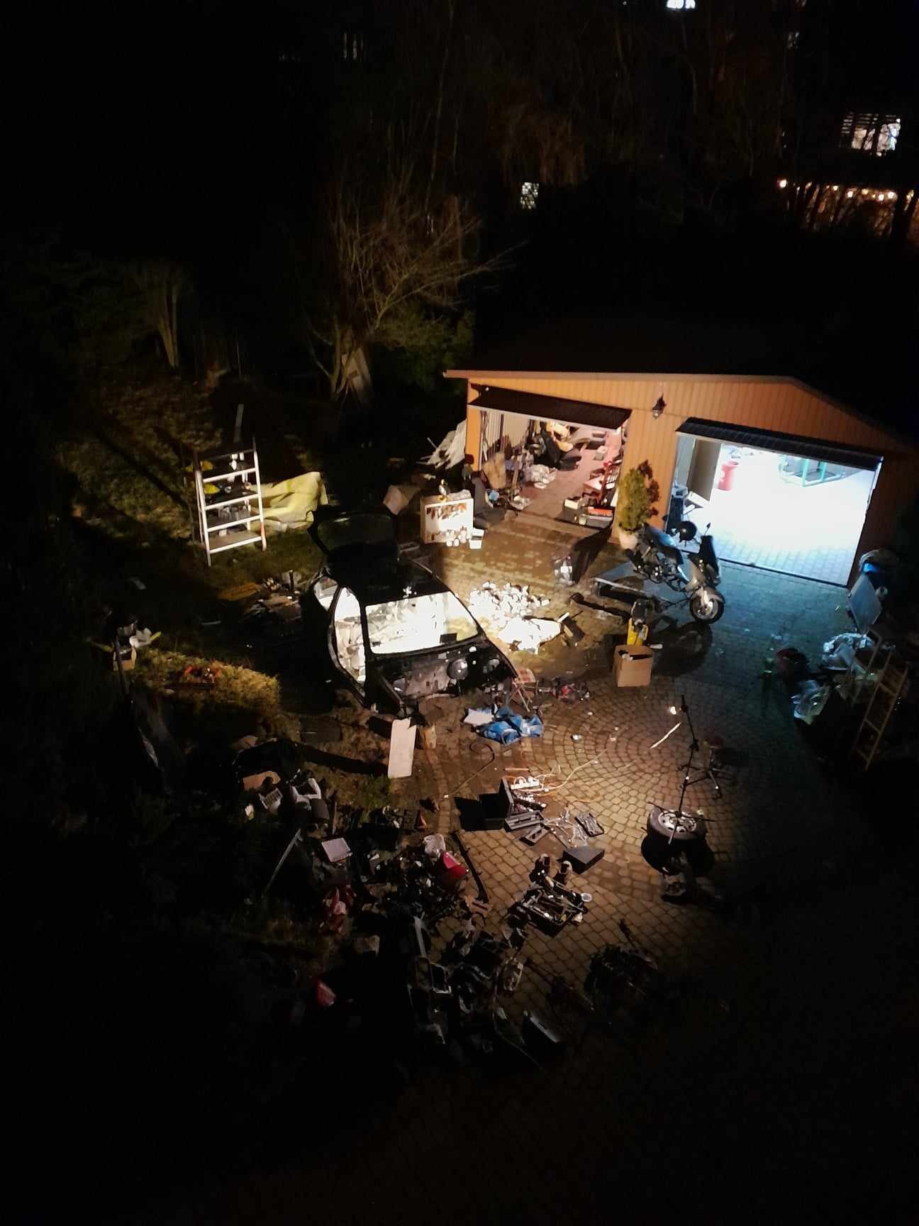 Nocne ujęcie z góry: rozmontowany szkielet samochodu na brukowanym podjeździe przed garażem, częściowo oświetlonym, obok zaparkowany motocykl i rozrzucone narzędzia.