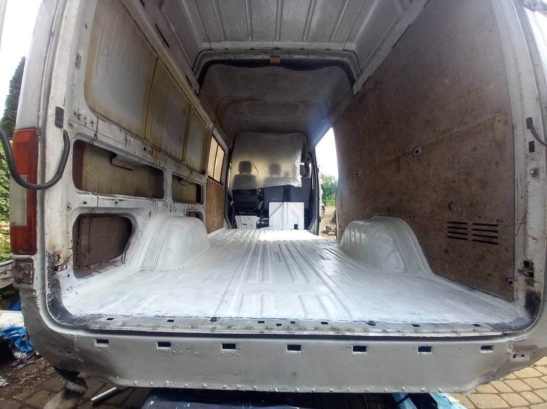 Wnętrze furgonetki w trakcie renowacji, widok z tyłu na odświeżoną, pomalowaną na biało podłogę, surowe ściany boczne bez tapicerki, widoczne ślady korozji i spawania na karoserii.