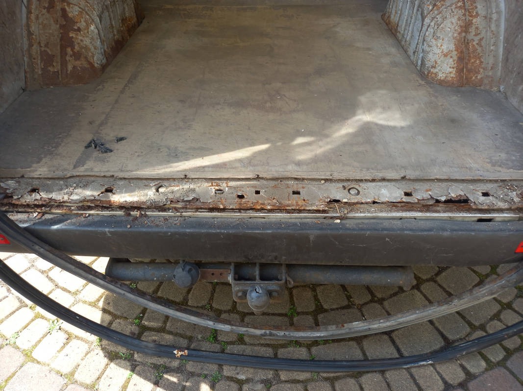 Wnętrze bagażnika samochodu dostawczego z widoczną rdzą na progach i elementach karoserii, hak holowniczy na tle kostki brukowej.