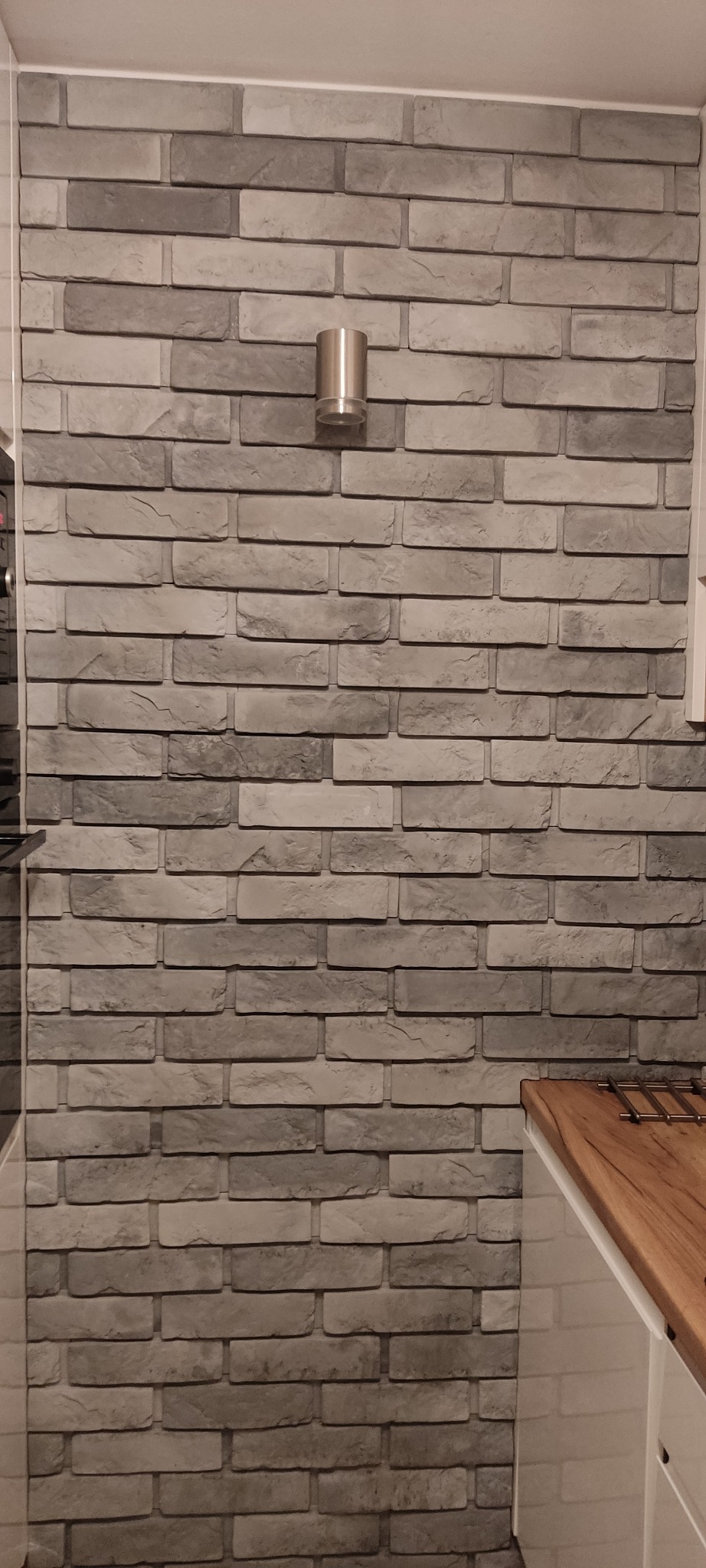 Ściana wykończona szarą cegłą dekoracyjną z oświetleniem punktowym, widoczny fragment blatu kuchennego.