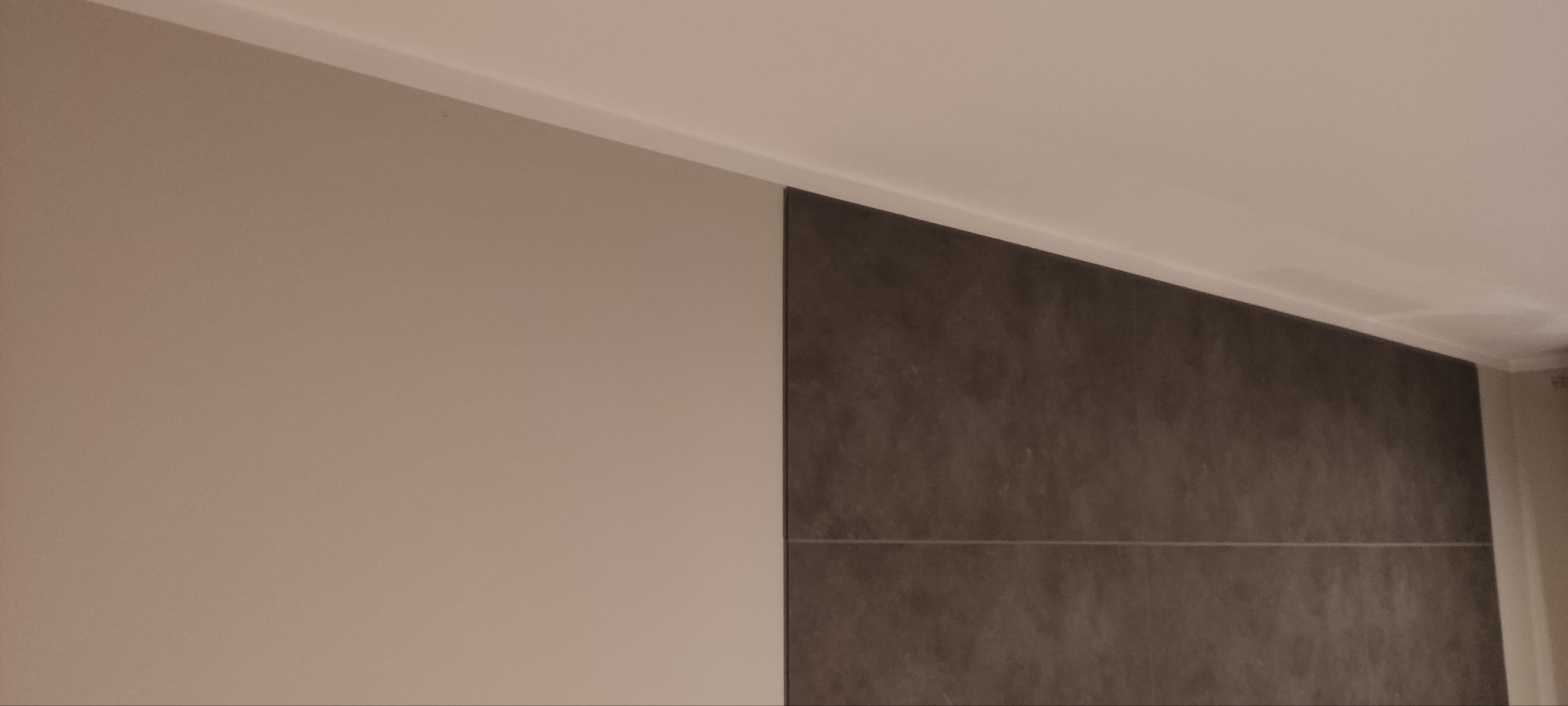 Fragment pomalowanej na beżowo ściany sąsiadującej z ciemnoszarą ścianą wykończoną płytkami imitującymi kamień, widoczne białe listwy przysufitowe.