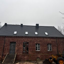 USŁUGI BUDOWLANE JAKUB KAMIŃSKI - Ceglany budynek z nowym, ciemnym dachem i czterema oknami połaciowymi, obok żółta koparka. Stare drewniane drzwi i gołe drzewa po bokach, w trakcie remontu.