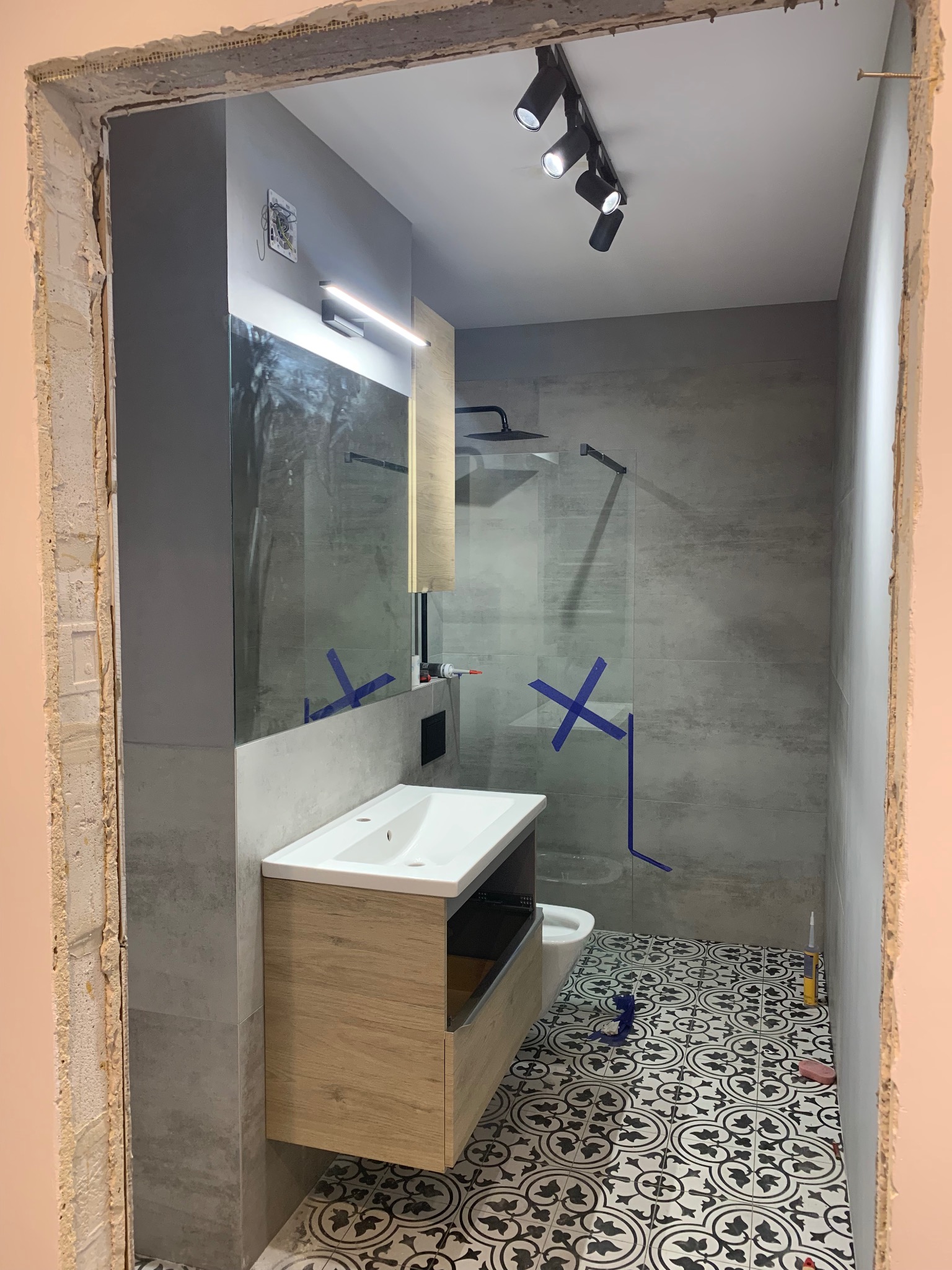 Wnętrze łazienki w trakcie wykańczania: umywalka z szafką z jasnego drewna, szklana kabina prysznicowa z deszczownicą, czarno-biała wzorzysta podłoga, ściany wyłożone szarymi płytkami, widoczne...