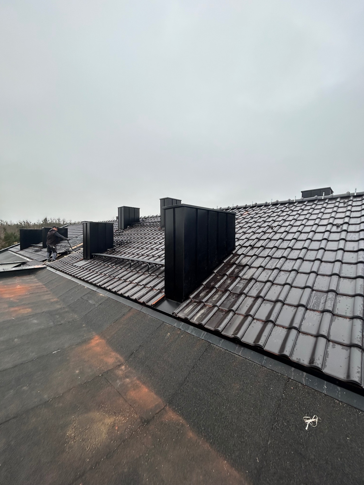 Pracownik montuje czarne kominy wentylacyjne na nowym, ciemnym dachu z dachówki ceramicznej w pochmurny dzień.