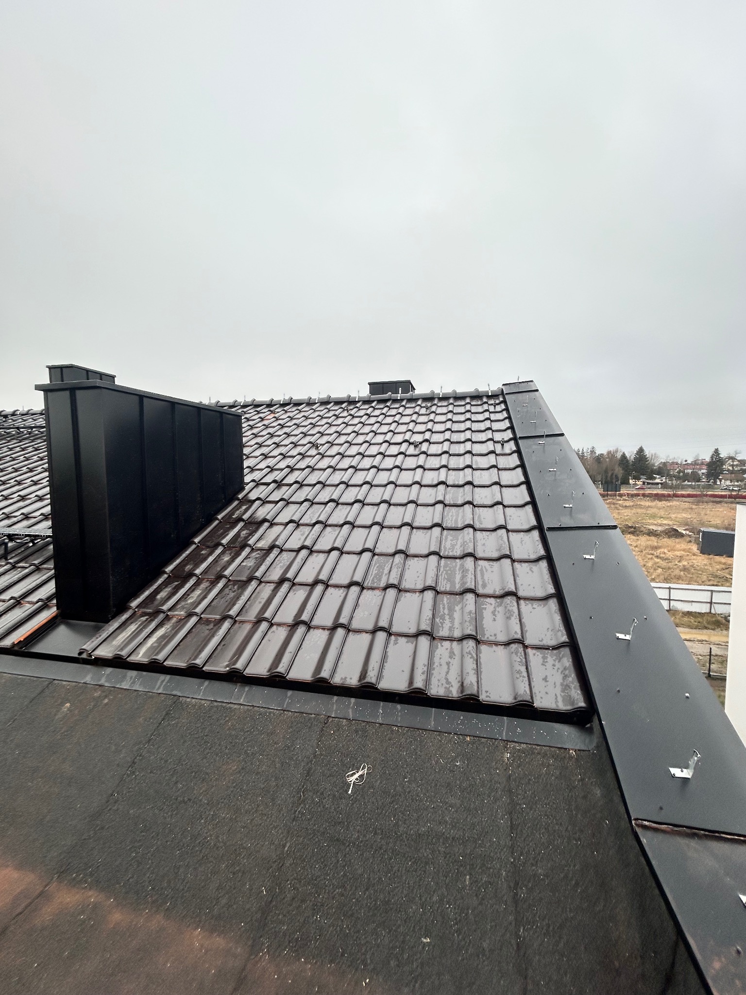 Widok z góry na fragment dachu pokrytego brązową dachówką ceramiczną, z czarną obróbką blacharską i kominem, w tle widoczne pole i zabudowania.