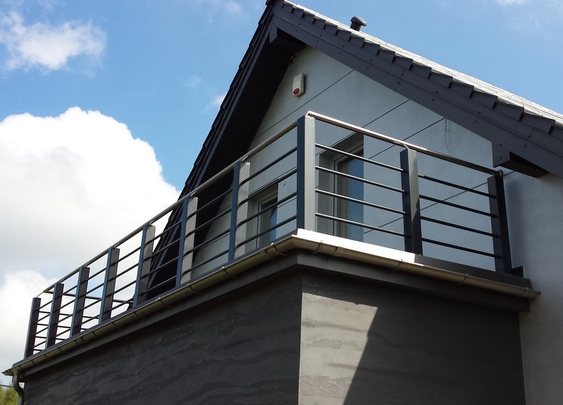 Balkon z nowoczesną, poziomą balustradą ze stali nierdzewnej w kolorze grafitowym, zamontowaną na budynku z szarą elewacją i ciemną dachówką, widok z dołu na tle błękitnego nieba z chmurami.