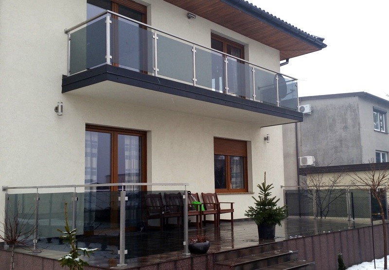 Nowoczesny dom z balkonem i tarasem zabezpieczonym szklanymi balustradami na metalowych słupkach. Widoczne drewniane okna i drzwi balkonowe oraz mała choinka w doniczce na tarasie.