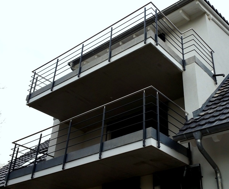 Nowoczesne, czarne balustrady z poziomych prętów na dwóch balkonach budynku z jasną elewacją i ciemnym dachem. Ujęcie z dołu.