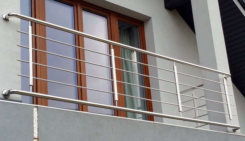 Balkon z nowoczesną, poziomą konstrukcją ze stali nierdzewnej, widok z dołu, na tle okna z brązową ramą i jasnoszarej elewacji.