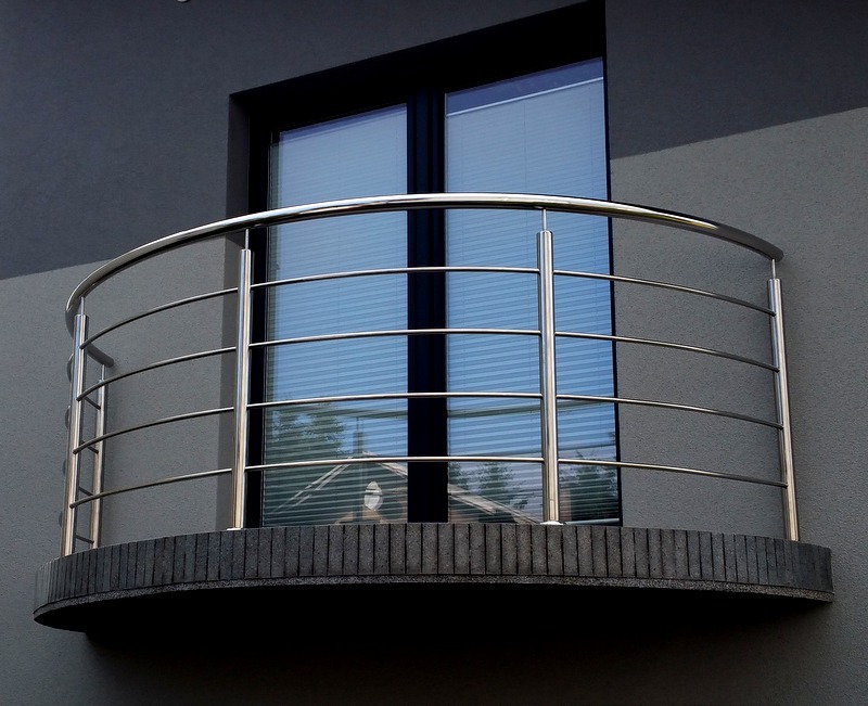 Krótki, półokrągły balkon z nowoczesną, metalową barierką na tle szarej fasady budynku i okna z żaluzjami.
