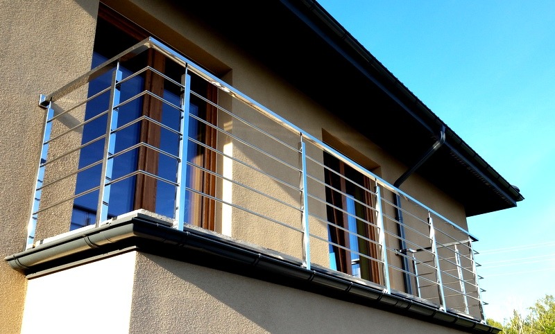 Balkon z nowoczesną, minimalistyczną balustradą ze stali nierdzewnej z cienkimi, poziomymi prętami, z widokiem na błękitne niebo i fragment dachu budynku.