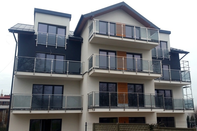 Trzypiętrowy budynek mieszkalny z balkonami zabezpieczonymi szklanymi taflami w stalowych ramach.