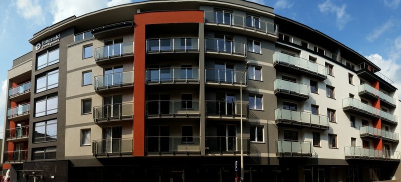 Nowoczesny budynek mieszkalny z balkonami zabezpieczonymi szklanymi barierkami, widok z dołu, błękitne niebo w tle.