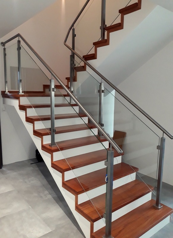 Schody z drewnianymi stopniami i szklaną balustradą z elementami ze stali nierdzewnej, widok z dołu na białe ściany i szarą podłogę.