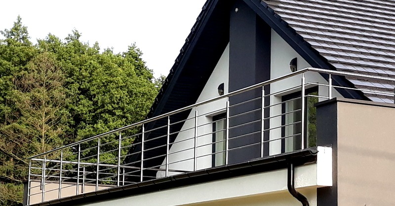 Balkon z nowoczesną balustradą nierdzewną, widok z zewnątrz na tle domu z czarnym dachem i zielenią drzew.