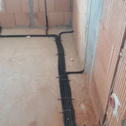 Wiązka czarnych, karbowanych rur z przewodami elektrycznymi, mocowana metalowymi uchwytami do surowej ściany z cegły dziurawki w Kosakowie, na etapie budowy.