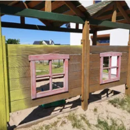 Drewniany domek zabaw dla dzieci z dwoma małymi okienkami w czerwonych ramach, częściowo pomalowany na żółto, stojący na piaszczystym placu zabaw.