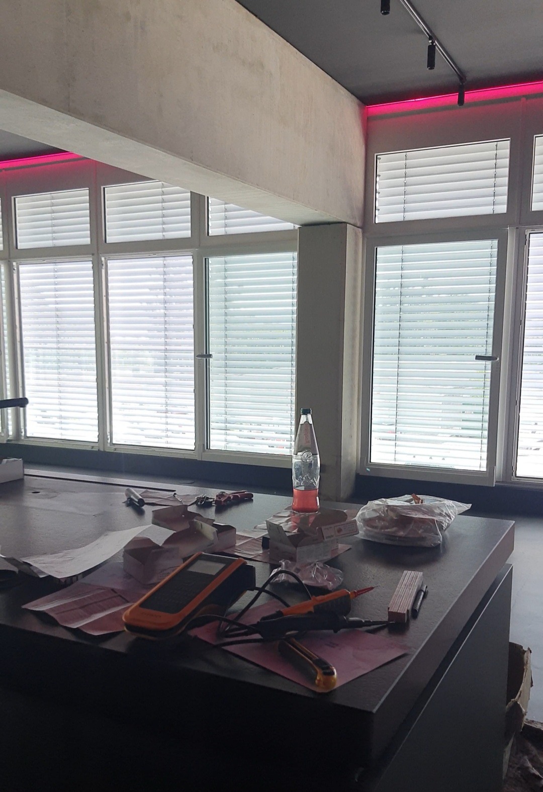 Multimetr, wkrętak, nóż i dokumentacja leżą na stole w pomieszczeniu z dużymi oknami i oświetleniem LED, sugerując przygotowania do prac elektrycznych.