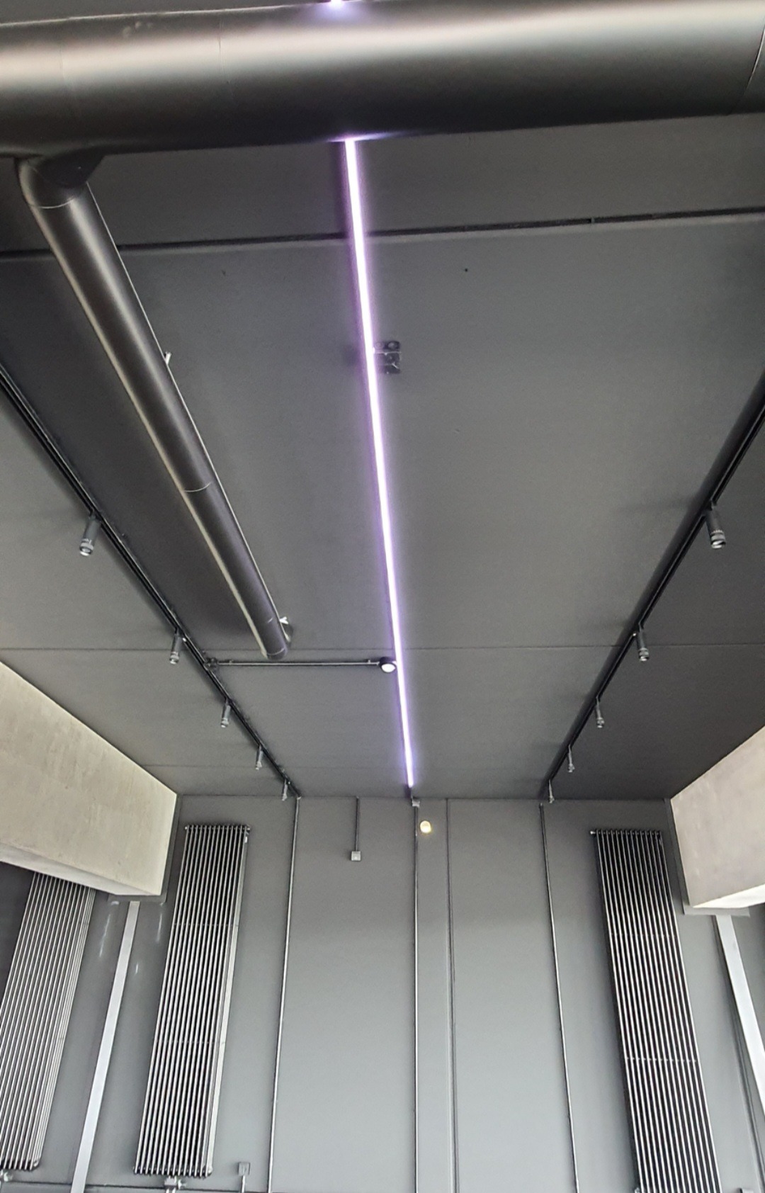 Szare wnętrze z widoczną instalacją oświetleniową: podwieszane reflektory punktowe na szynoprzewodach oraz liniowa lampa LED, rury wentylacyjne pod sufitem, grzejniki panelowe na ścianach.