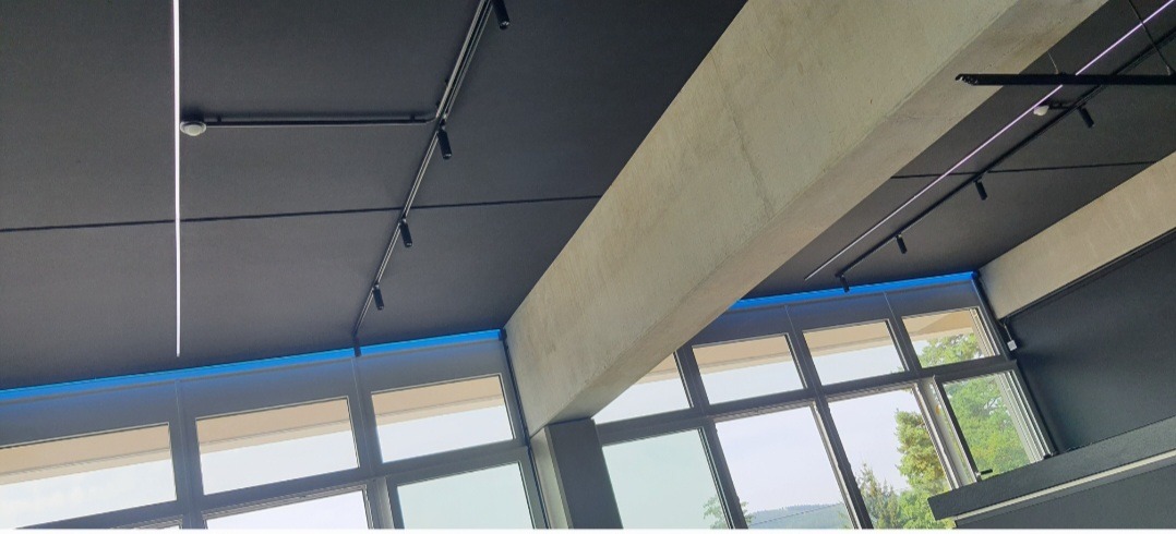 Nowoczesne oświetlenie sufitowe z szynoprzewodami i reflektorami punktowymi w pomieszczeniu z dużymi oknami i betonową belką stropową.