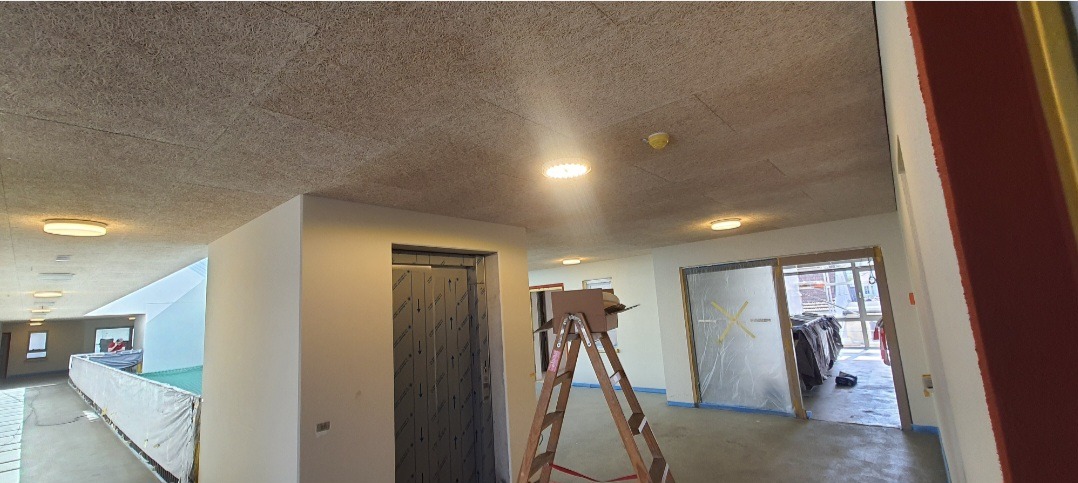 Korytarz w trakcie prac wykończeniowych: sufit z płyty OSB, zamontowane okrągłe lampy, zabezpieczone folią drzwi i winda, drabina w centrum kadru.