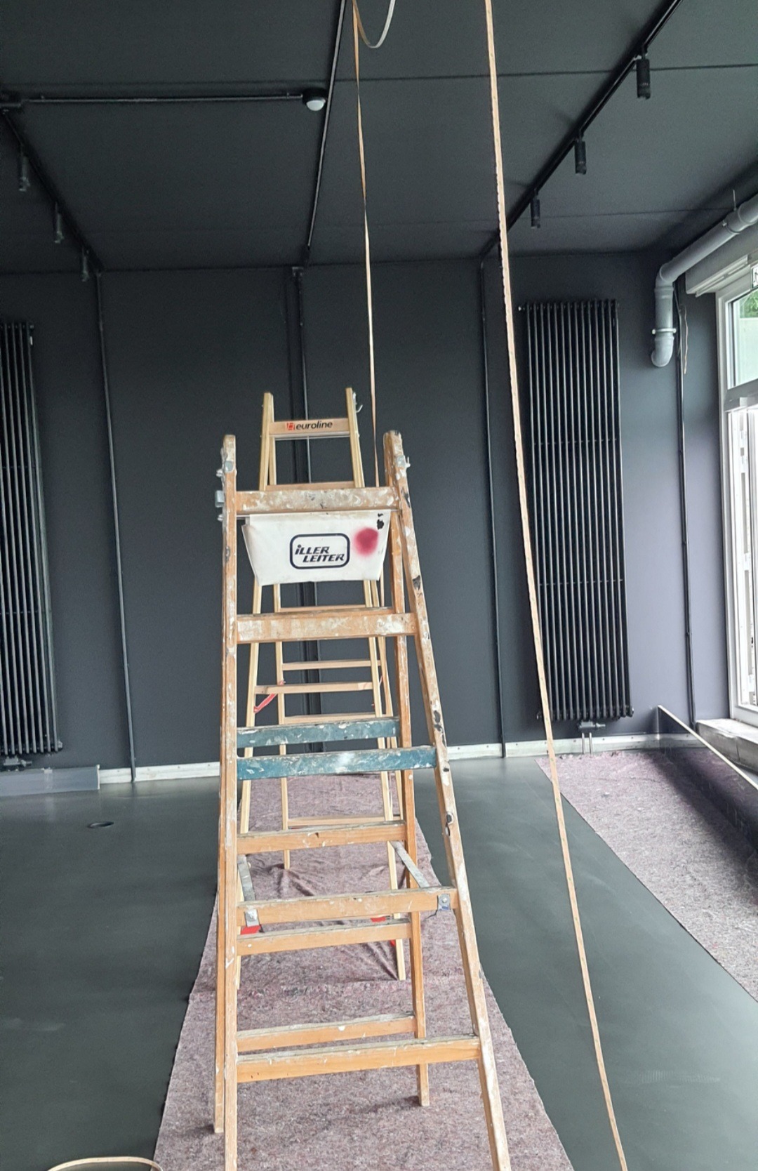 Przygotowania do malowania wnętrza: drewniana drabina z wiaderkiem z farbą na tle ciemnoszarej ściany i grzejnika, zabezpieczona podłoga.