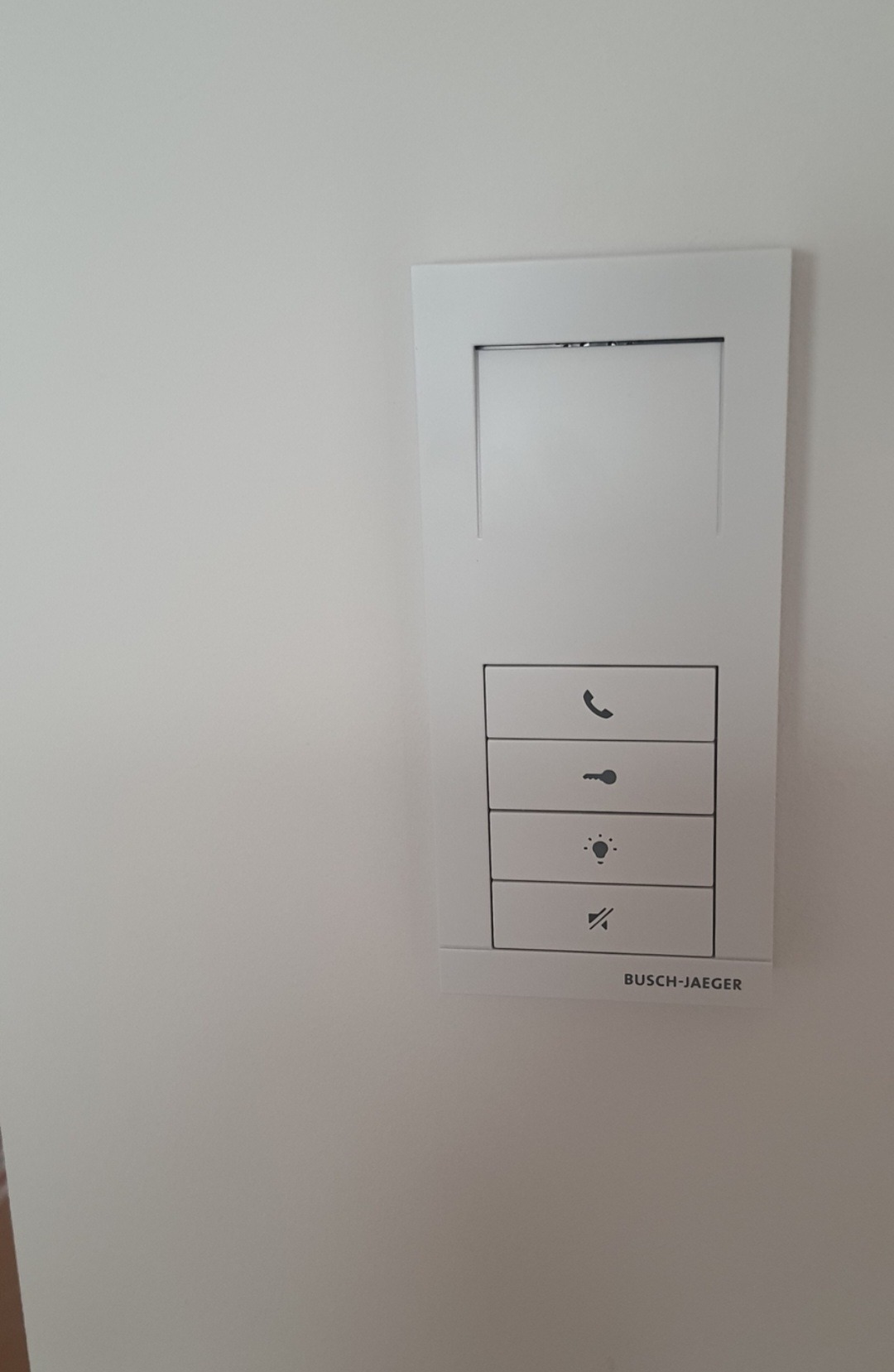 Panel sterowania inteligentnego domu Busch-Jaeger z przyciskami do sterowania oświetleniem, domofonem i innymi funkcjami, zamontowany na białej ścianie.