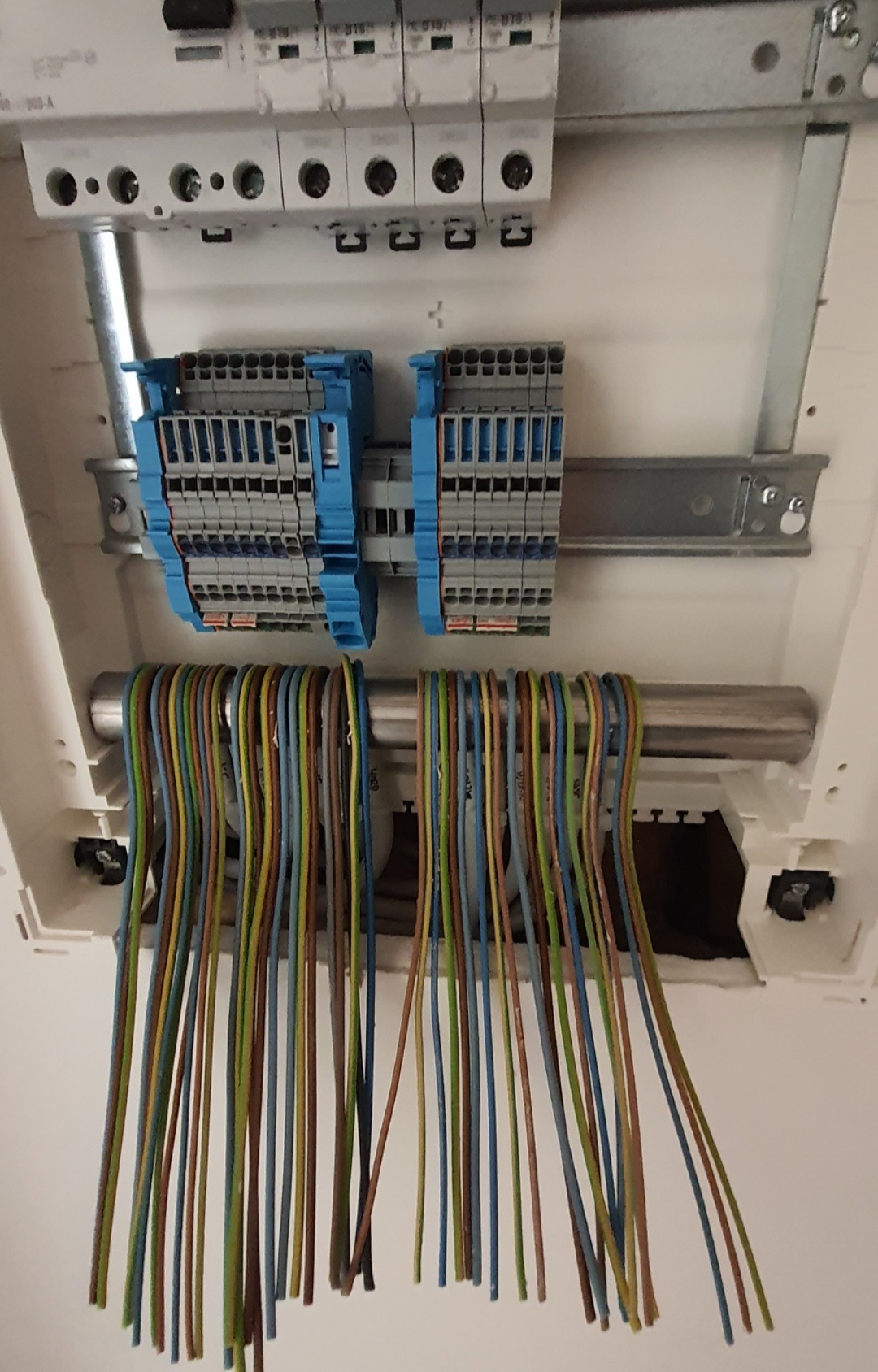 Rozdzielnica elektryczna z podłączonymi przewodami o różnych kolorach, zamocowana na białej ścianie, widoczne zabezpieczenia i listwy zaciskowe.