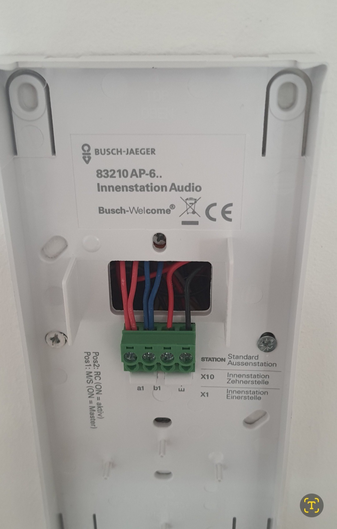 Widok z tyłu białej stacji wewnętrznej audio Busch-Jaeger 83210 AP-6 z podłączonymi przewodami: czerwonym, niebieskim i czarnym, wpiętymi w zieloną listwę zaciskową.