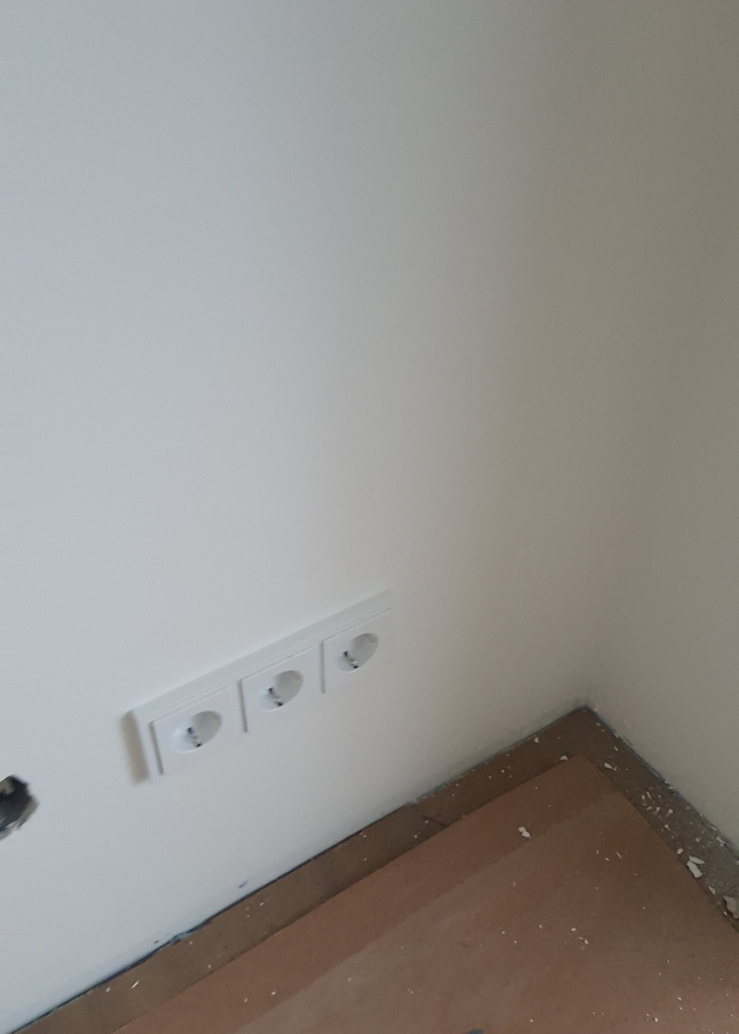 Potrójne, białe gniazdko elektryczne zamontowane na świeżo pomalowanej, białej ścianie w rogu pomieszczenia; widoczne ślady po remoncie na podłodze.