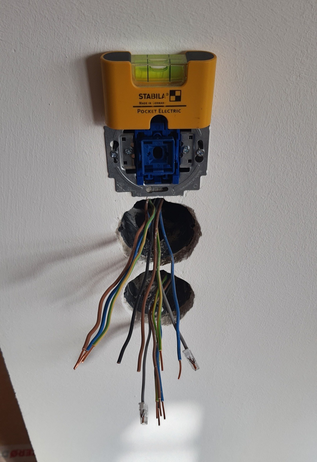 Żółta poziomica Stabila Pocket Electric przymocowana do puszki elektrycznej w ścianie, z widocznymi przewodami o różnych kolorach wychodzącymi z dwóch otworów pod puszką.