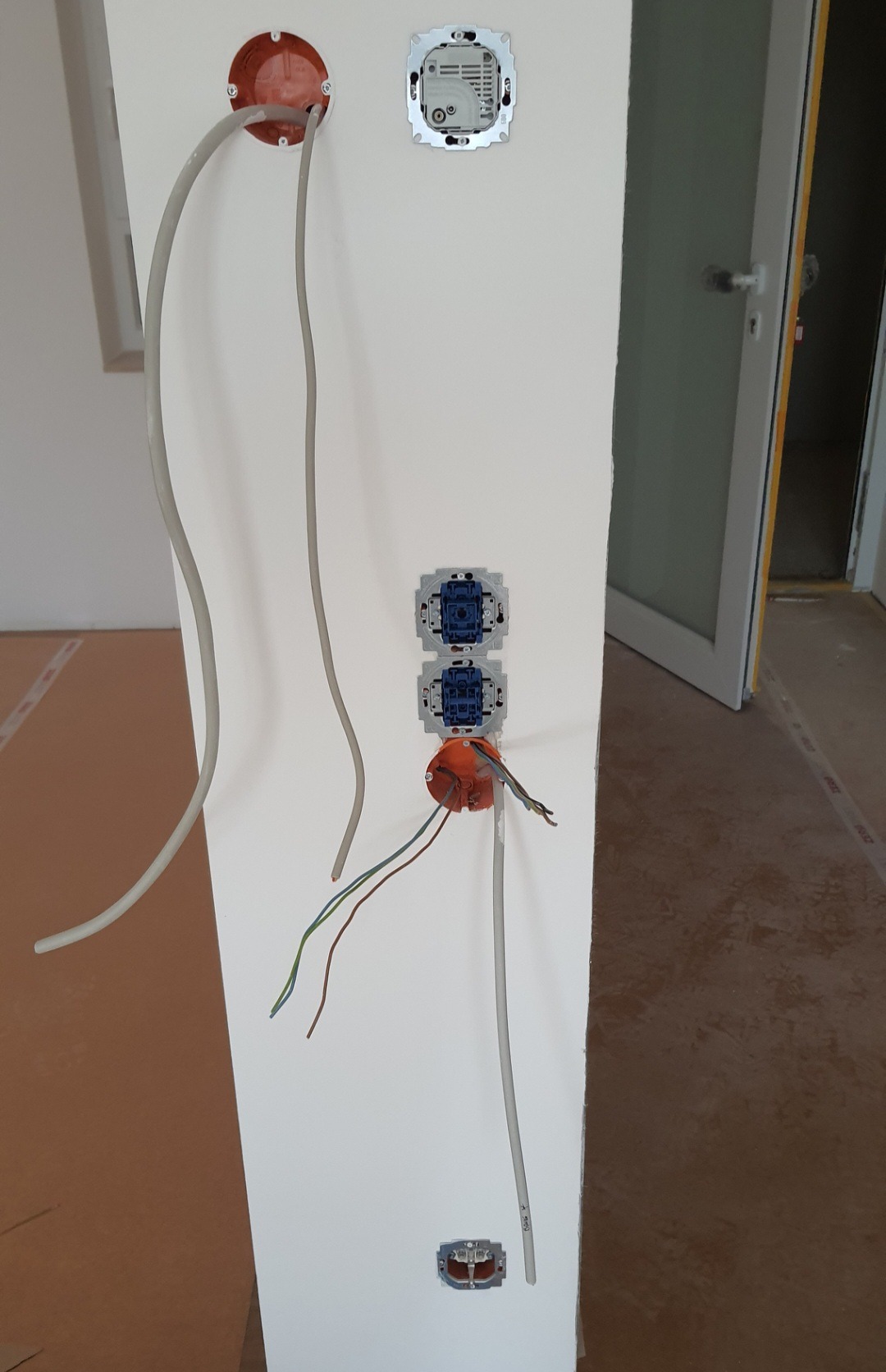 Ściana w trakcie prac elektrycznych: widoczne puszki instalacyjne z wystającymi przewodami, gniazda bez obudowy oraz kable prowadzone po ścianie.