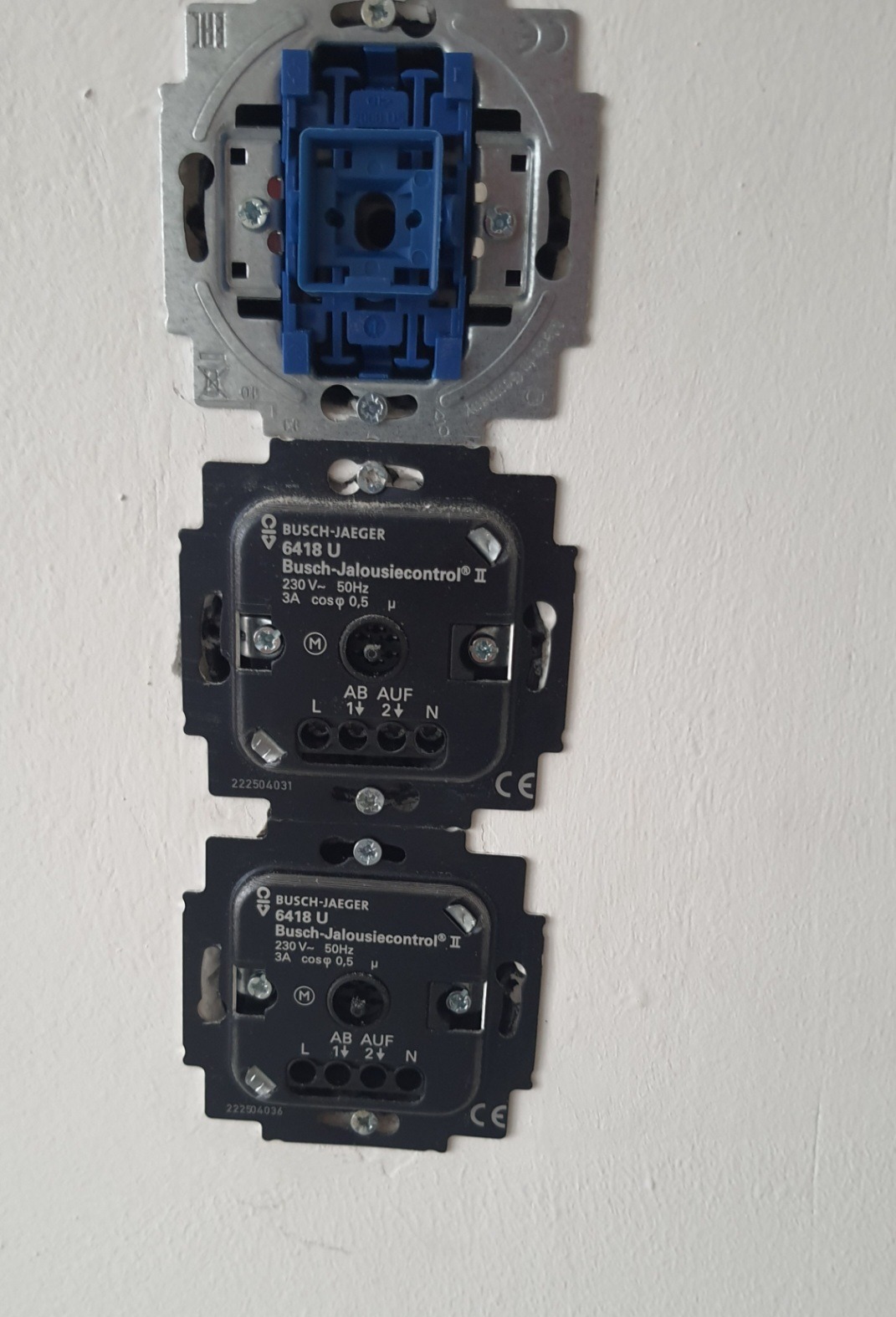 Widok z bliska na zamontowane, ale jeszcze niekompletne, dwa czarne moduły sterowania roletami Busch-Jaeger 6418 U, przymocowane śrubami do ściany, z widoczną metalową ramką montażową i niebieskim...