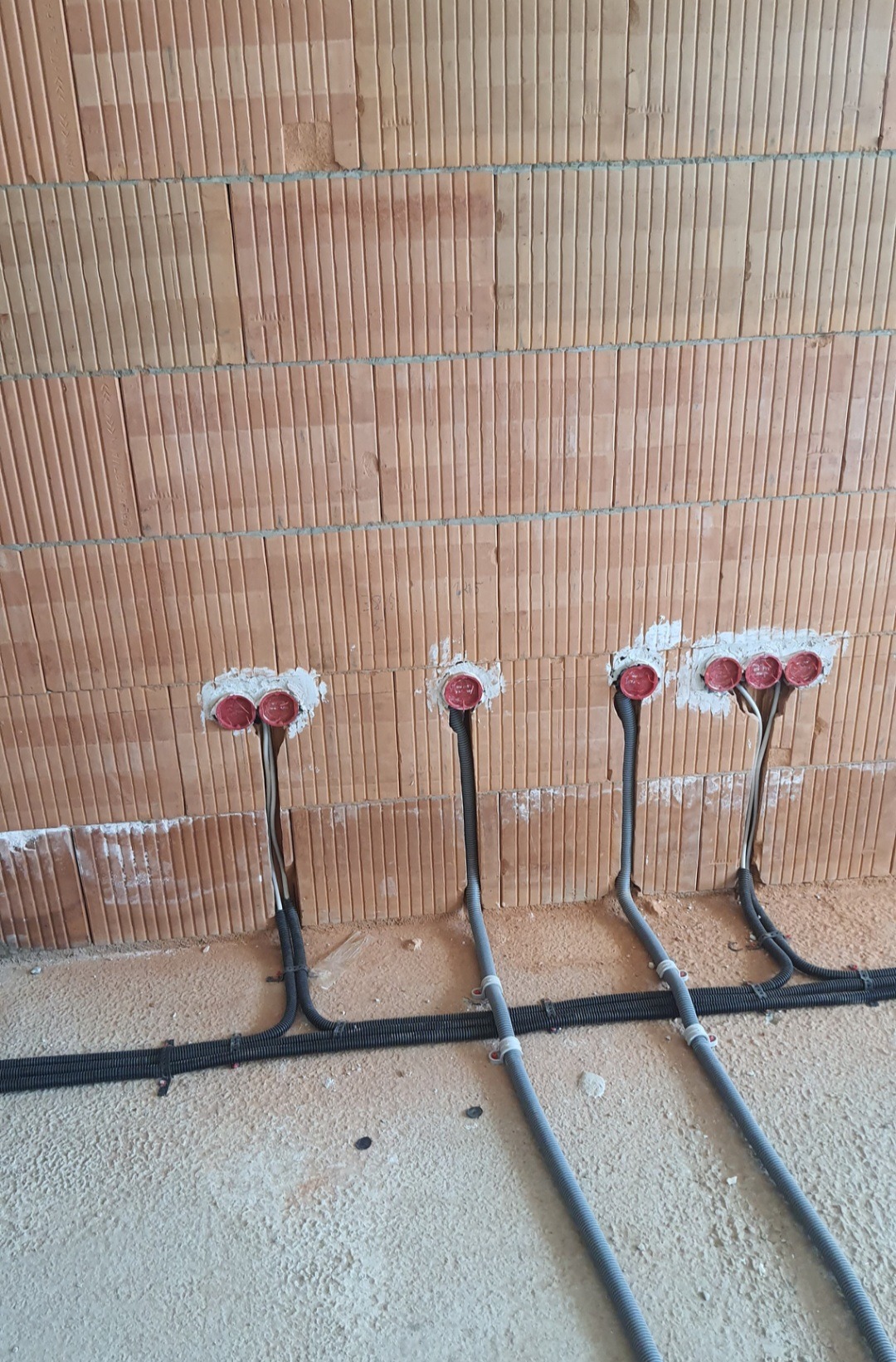 Instalacja elektryczna w trakcie montażu w ścianie z czerwonej cegły, widoczne puszki instalacyjne z przewodami i peszle na podłodze.