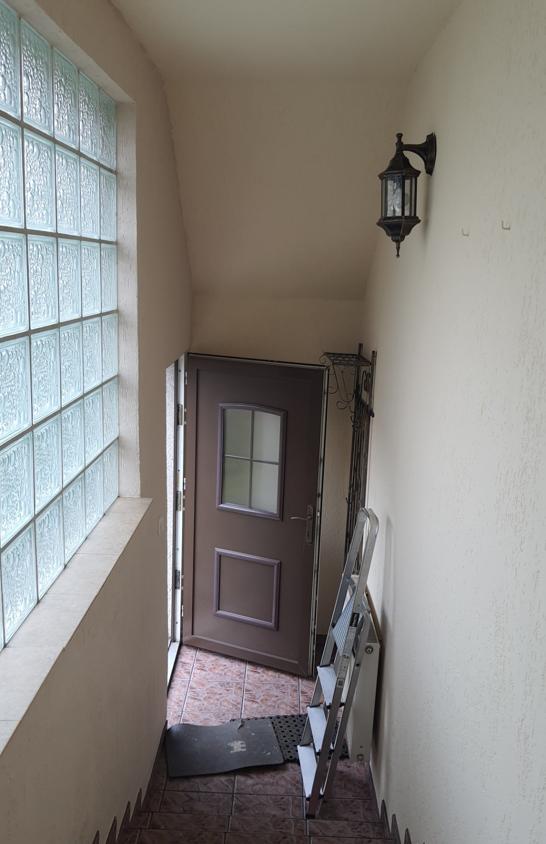 Korytarz wejściowy z oknem z luksferów, brązowymi drzwiami i metalową lampą ścienną, widoczna drabina aluminiowa.