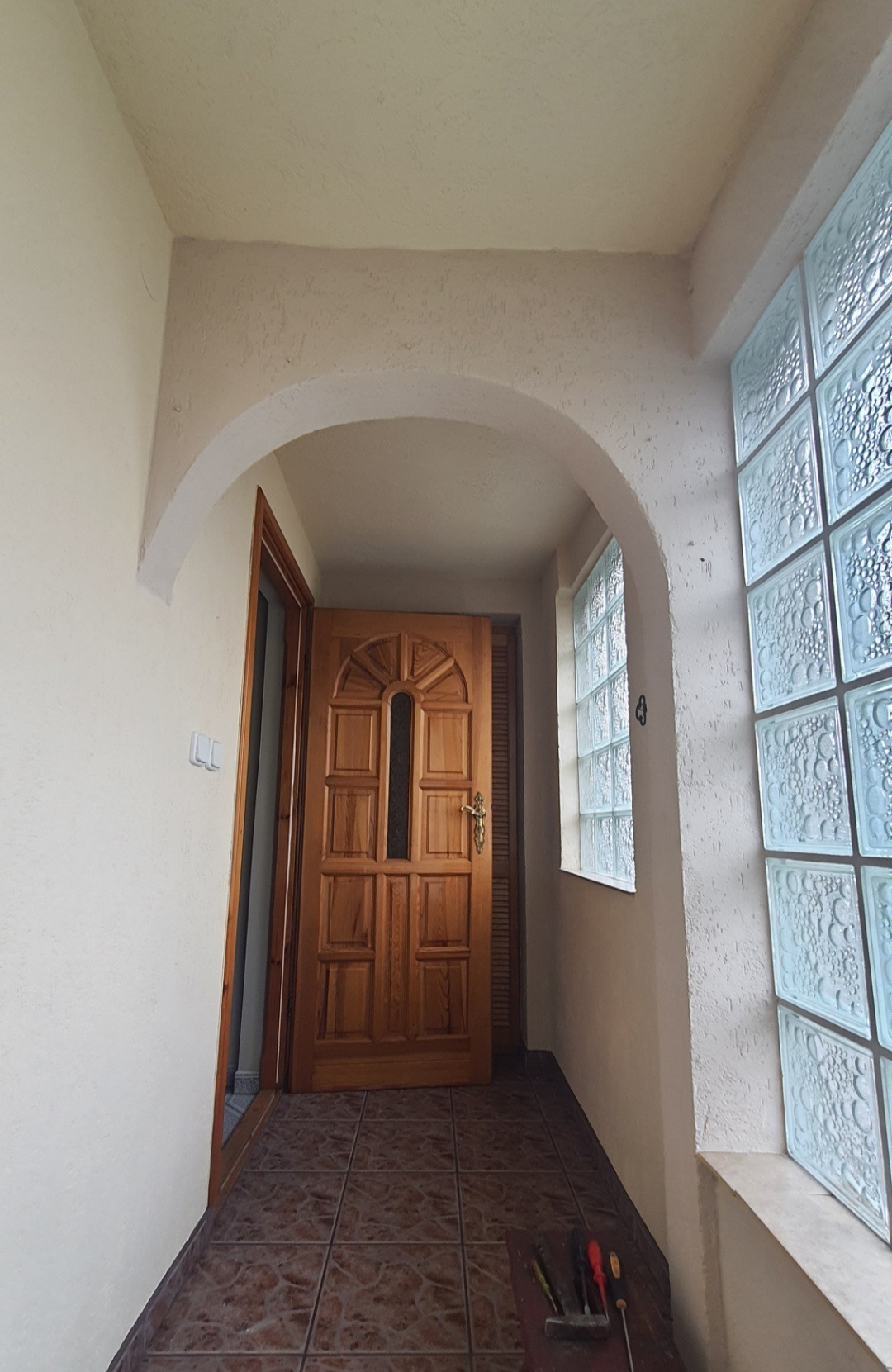 Wnętrze przedpokoju z drewnianymi drzwiami wejściowymi, oknem z luksferami i terakotą na podłodze. Widoczne ślady świeżo zakończonych prac malarskich na ścianach i suficie.