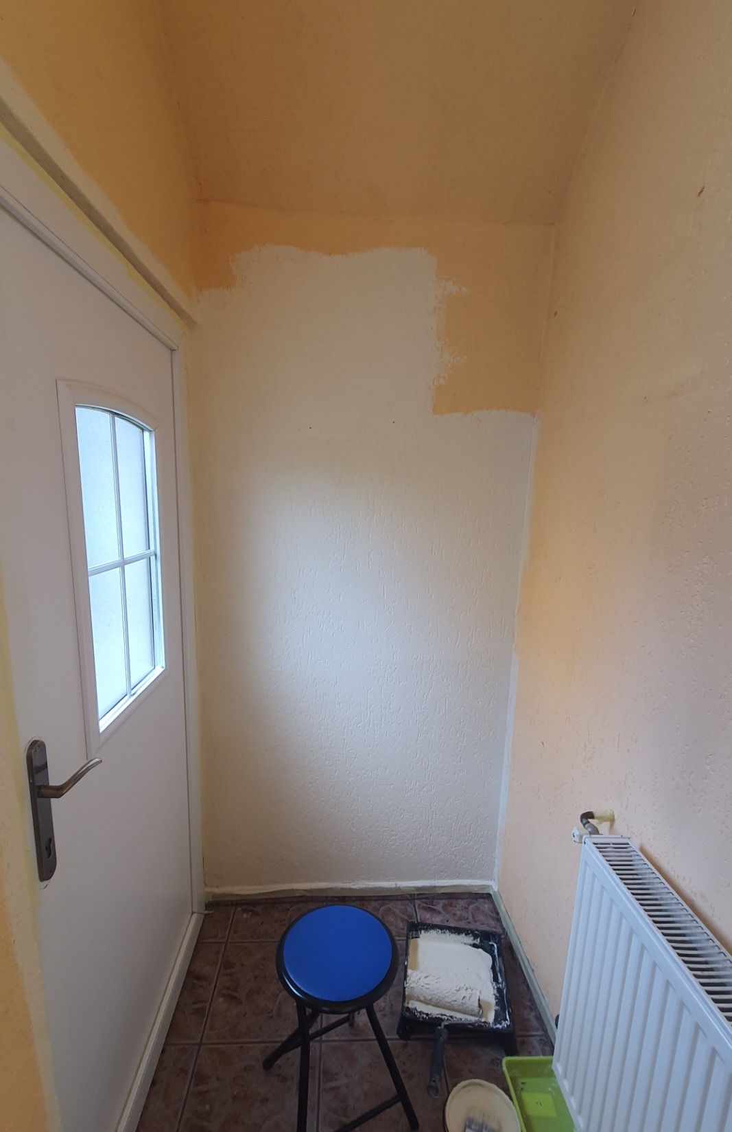 Fragment ściany w trakcie malowania na biało, widoczny wałek w kuwecie z farbą, niebieski taboret i grzejnik.