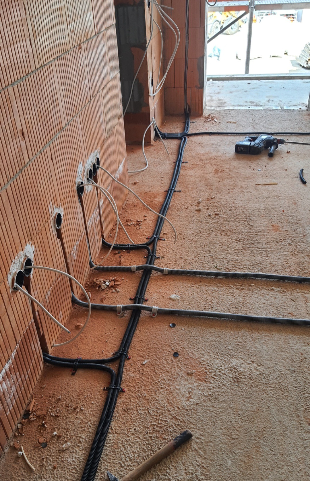 Wewnętrzne okablowanie elektryczne w surowym stanie budynku, widoczne przewody wystające ze ścian z pustaków ceramicznych, rury karbowane prowadzone po podłodze, w tle wiertarka akumulatorowa.