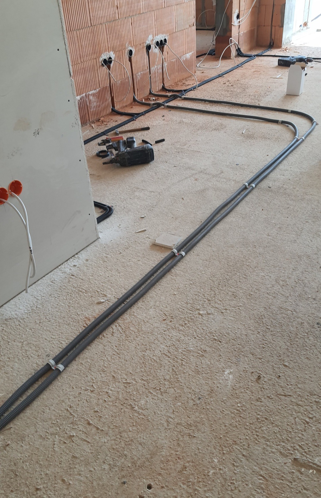 Wnętrze w trakcie prac wykończeniowych: widoczne kable elektryczne w peszlach ułożone na podłodze, puszki instalacyjne na ścianie z karton-gipsu oraz narzędzia budowlane w tle, ceglana ściana...