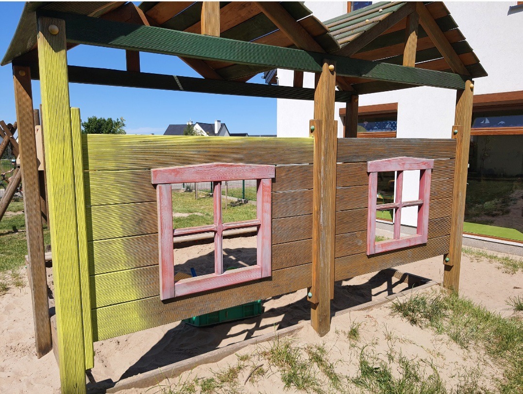 Drewniany domek zabaw dla dzieci z dwoma małymi okienkami w czerwonych ramach, częściowo pomalowany na żółto, stojący na piaszczystym placu zabaw.