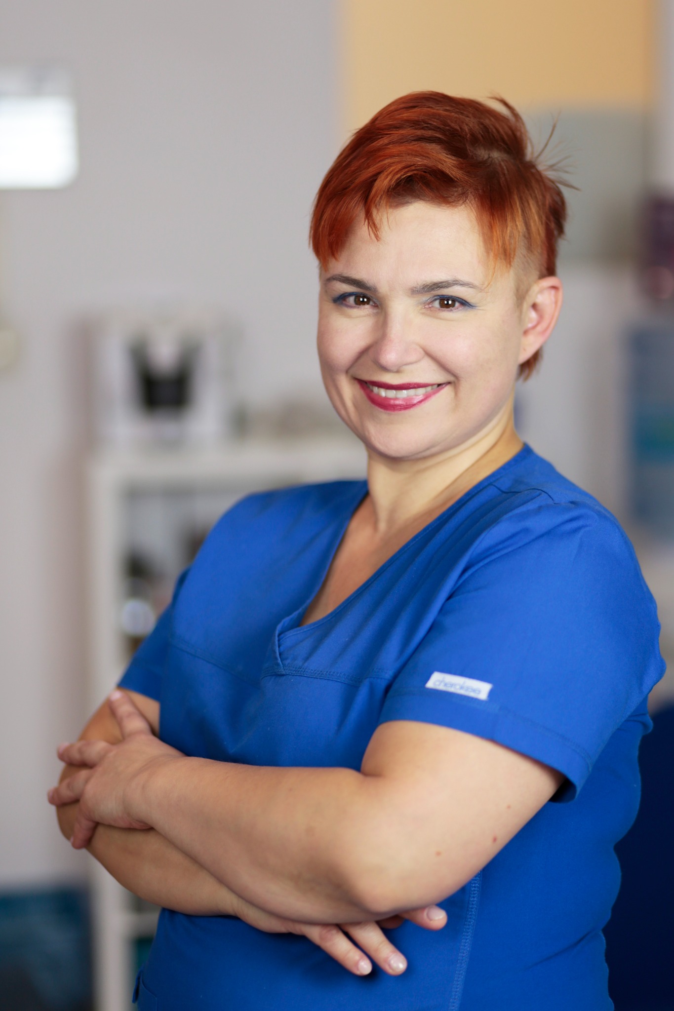 Uśmiechnięta kobieta w niebieskim uniformie medycznym, z krótkimi rudymi włosami, stoi na tle jasnego wnętrza gabinetu. Ręce skrzyżowane na piersiach.