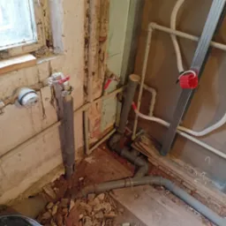 Fragment instalacji hydraulicznej w trakcie przeróbki: szare rury PCV, metalowe profile do zabudowy, fragment okna z widocznymi śladami wilgoci i zniszczeń na ścianie.