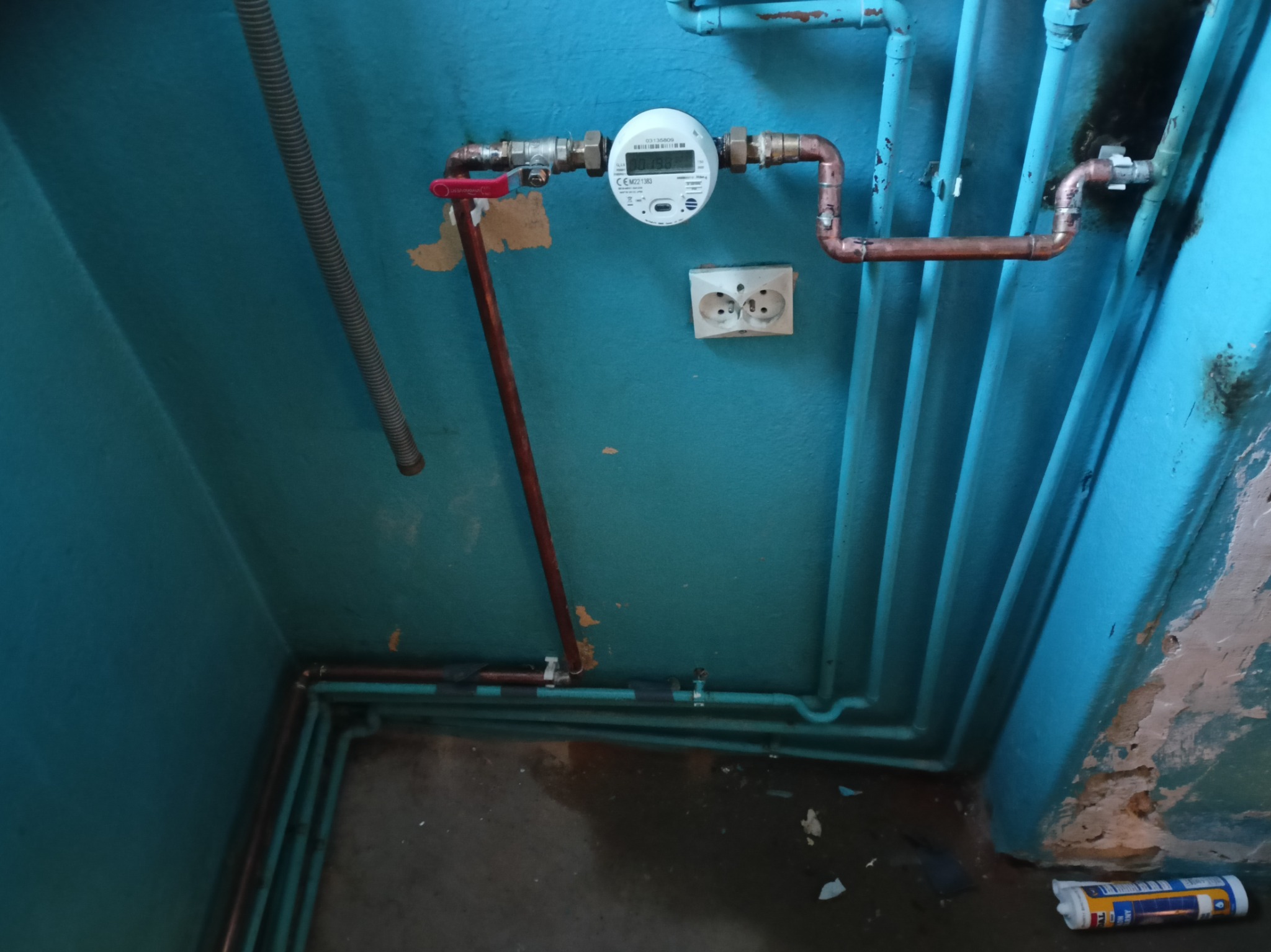 Wodomierz z zaworem odcinającym i instalacją miedzianą na tle pomalowanej na niebiesko ściany z widocznymi starymi rurami i gniazdkiem elektrycznym.